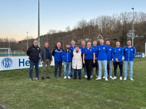 Eine Gruppe von elf Personen, teilweise in Vereinskleidung des SV Osterburken (blaue Trainingsjacken), steht gemeinsam auf einem Rasensportplatz vor einer Werbebande mit dem Vereinslogo. Im Hintergrund sind Bäume und ein Flutlichtmast zu sehen.